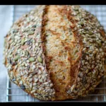 Freshly baked seeded multigrain sourdough bread loaf with a golden crust and hearty texture, displayed on a rustic wooden table.