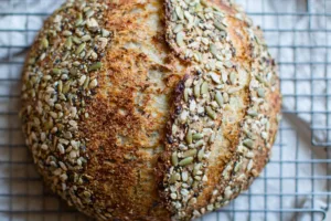 Seeded Multigrain Sourdough Bread Recipe Freshly baked seeded multigrain sourdough bread loaf with a golden crust and hearty texture, displayed on a rustic wooden table.