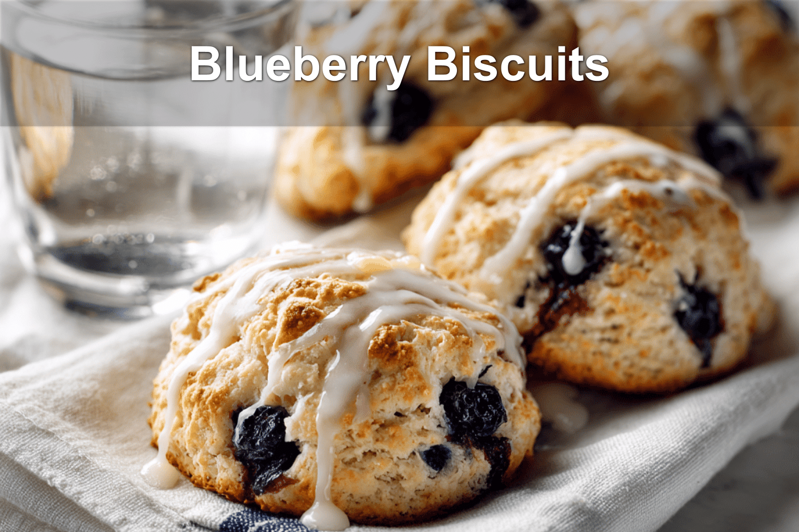 Golden, fluffy blueberry biscuits topped with glaze on a clean napkin with water on the side.