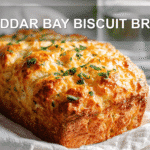 CHEDDAR BAY BISCUIT BREAD loaf on white napkin, warm light, golden crust, glass of water beside.