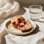 Plated raspberry sourdough muffin with honey drizzle beside water glass on white napkin.