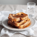 Plated sourdough French toast garnished with syrup and cinnamon, paired with a glass of water.