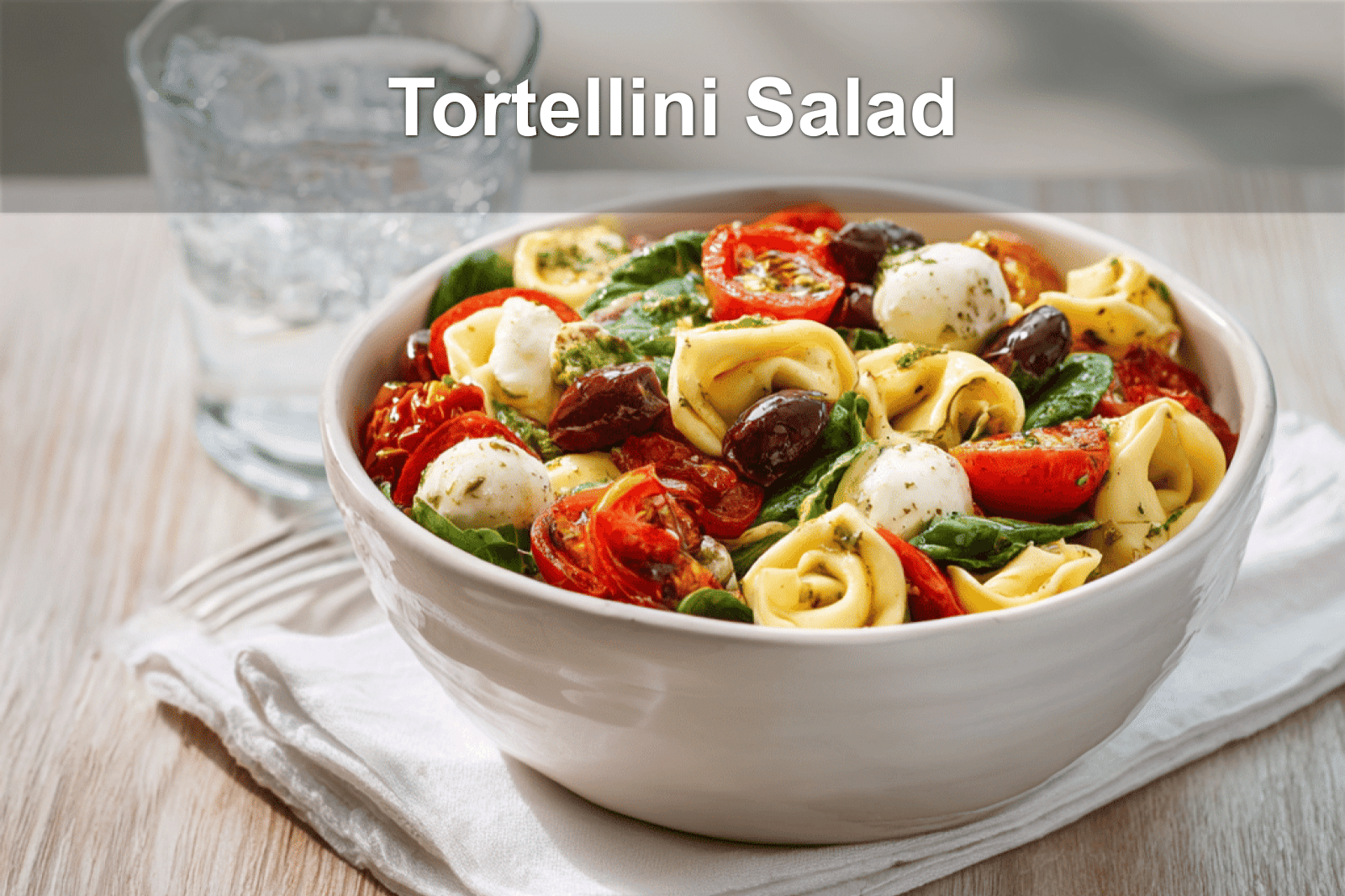 Vibrant tortellini pasta salad with fresh ingredients served in a white bowl, glass of water on the side.